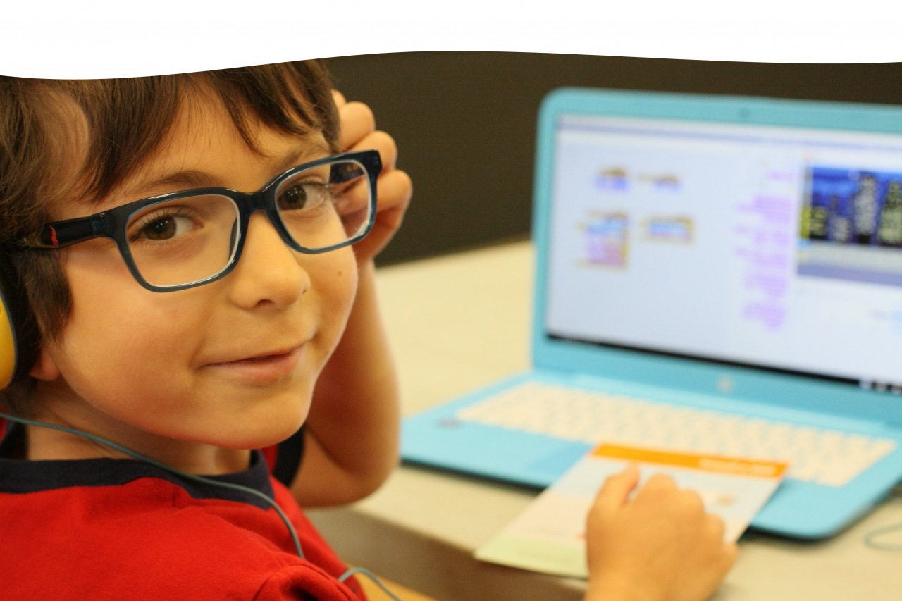 Young boy in red shirt and glasses smiles at the camera. He is sitting in front of a computer that has a Scratch Coding program running.